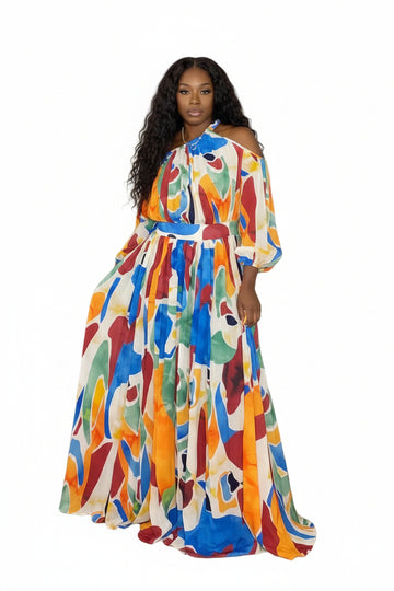 A colorful off-the-shoulder maxi dress with an abstract, painterly pattern in blue, orange, green, red, and yellow on a white base, styled with a matching waist belt and shown against a green artificial hedge background.